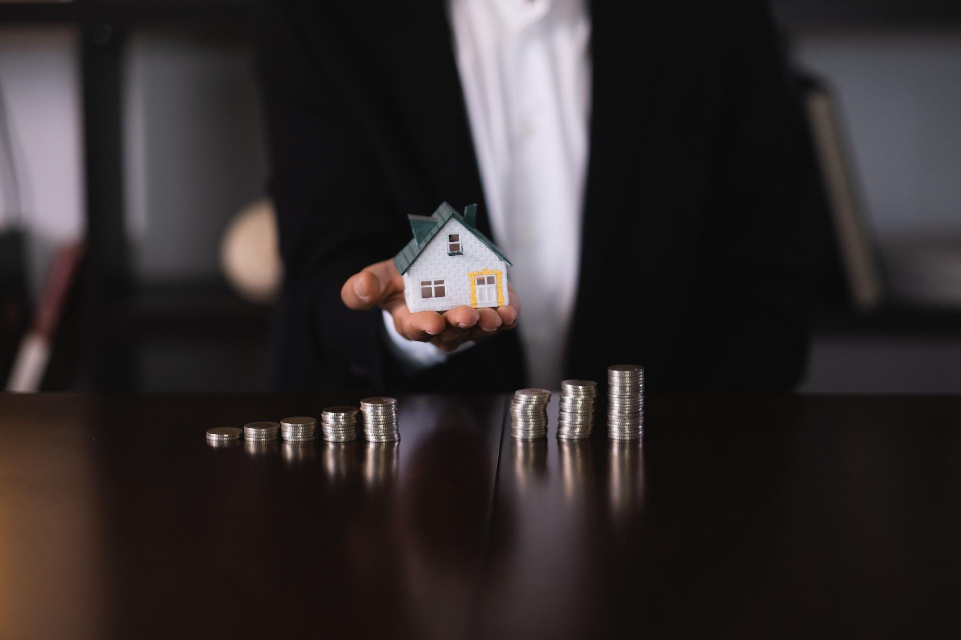 Real estate agent holding a model house with increasing stacks of coins on a table